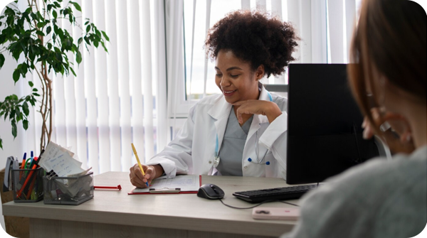 Descrição: Fotografia que mostra um cenário de consulta médica. A dra. atende uma pessoa (que tem cabelos longos e está de costas) e prescreve algo no papel. Ela tem uma expressão feliz e sorri. O consultório é iluminado, tem janelas grandes e cortina de persiana. A mesa onde a médica se encontra tem um notebook e porta-lápis. No canto esquerdo, aparece um vaso de planta.